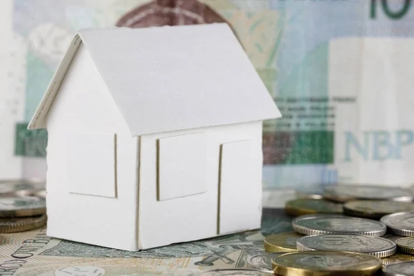 A model of a detached house made of white paper stands on Polish Zloty banknotes and coins which can mean real estate, mortgage or just a paper toy or other meaning of real estate