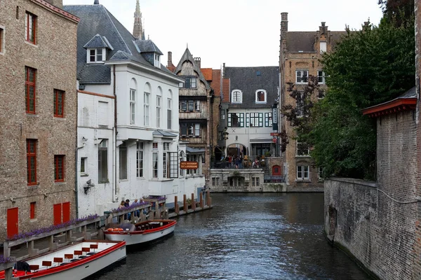 Bruges, Belçika - 8 Eylül 2022: Kanalın yanında tuğla binalar var, yanında insanların olduğu küçük bir iskele var, iki tekne turtayla demirlenmiş.