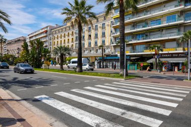 Nice, Fransa - 21 Nisan 2023: Güneşli bir günde gezinti güvertesi boyunca apartman binaları ve oteller görüldü. Ayrıca geniş bir cadde var, yaya geçidi görünüyor ve şu anda çok az trafik var. 