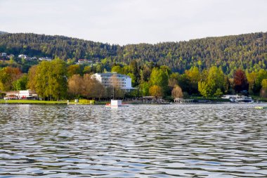 Velden Am Worthersee, Avusturya - 4 Mayıs 2023: Güzel doğanın yakınlarındaki evlere doğru göl manzarası burayı büyük bir tatil beldesi haline getiriyor