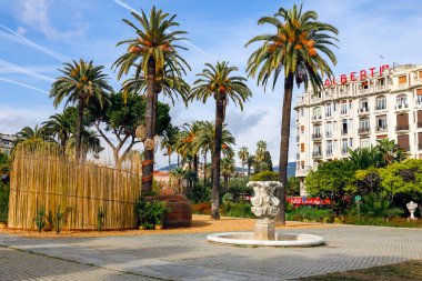 Güzel, Fransa - 23 Nisan 2023: Şehir parkındaki manzara palmiye ağaçlarını gösteriyor, küçük bir çeşme, biraz ileride büyük bir otel binasının parçası var.