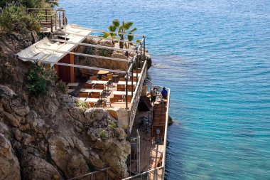 Nice, Fransa - 28 Nisan 2023: Açık hava deniz önü restoranı Akdeniz 'in hemen üzerindeki kayalık bir uçurumun üzerinde yer almaktadır. Bu restoranın masaları ve sandalyeleri sizi bekliyor.