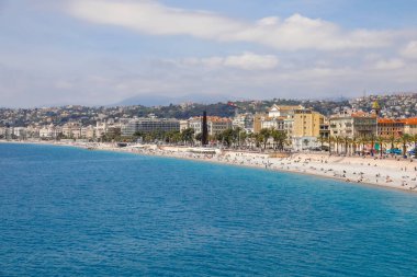 Nice, Fransa - 28 Nisan 2023 Akdeniz kıyısı boyunca ve şehirdeki binalar boyunca bir plaj. Sahilde deniz kenarındaki cazibe ve manzaranın tadını çıkaran insanlar var..