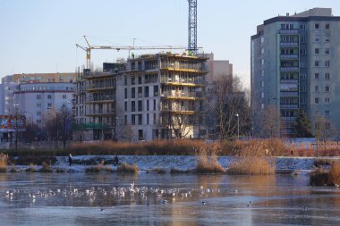 Warsaw, Poland - January 8, 2024: A new block of flats under construction, seen in winter behind a frozen lake in the Goclaw housing estate in Praga-Poludnie clipart