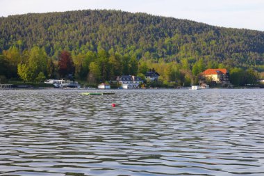 Velden Am Worthersee, Avusturya - 4 Mayıs 2023: Gölün karşısındaki ağaçlık tepeler ve güzel kırsal kesimin merkezindeki evler ile burası tatil için harika bir yer..