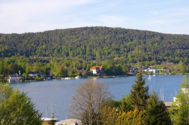 Velden Am Worthersee, Avusturya - 4 Mayıs 2023: Gölün karşısındaki ağaçlık tepeler ve güzel kırsal kesimin merkezindeki evler ile burası tatil için harika bir yer..