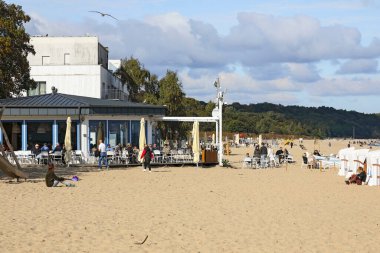 Sopot, Polonya - 6 Ekim 2024: Kumlu plajın kenarındaki açık hava bölümü olan restoran. Güneşli sonbahar gününün tadını çıkaran insanlar var..
