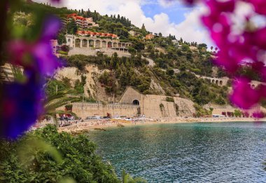 Fransa 'nın güneyinde Villefranche sur Mer ile Fransız Riviera sahili manzarası, bougainvillea çiçekleri ve palmiye ağaçları tarafından çerçevelenmiştir.