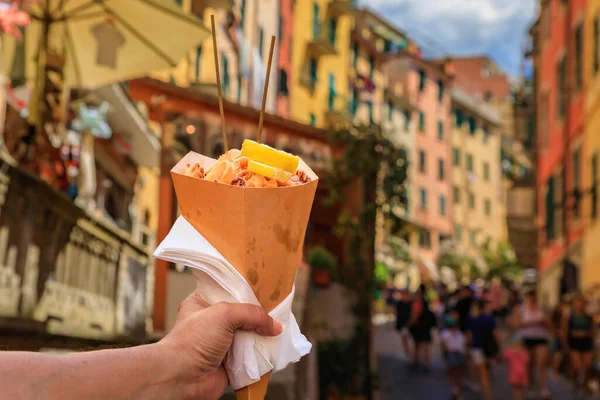 stock image Hand holding a cone of fritto misto, Italian street food mix of fried calamari and shrimp with colorful houses in Riomaggiore, Cinque Terre Italy