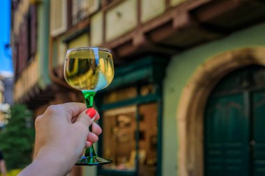 Açık hava restoranında bir bardak Gewurztraminer şarabı ile bir kadının eli, Colmar France, Alsace Wine Route köyündeki yarım tahtalı evleri bulanıklaştırdı.