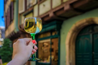 Açık hava restoranında bir bardak Gewurztraminer şarabı ile bir kadının eli, Colmar France, Alsace Wine Route köyündeki yarım tahtalı evleri bulanıklaştırdı.