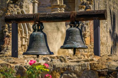 San Juan Capistrano, USA - August 6, 2023: Two weathered bells hang from an old wooden beam in front of a stone wall, Mission San Juan Capistrano clipart