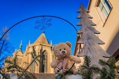 A teddy bear sits in front of a decorative archway at the Christmas Market with the spire of the Colmar Cathedral visible in the background, France clipart