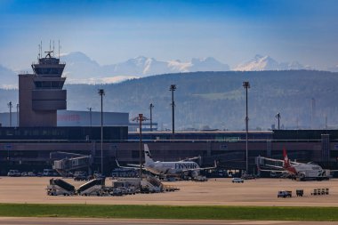 Zürih, İsviçre - 13 Nisan 2024: Kontrol kulesine sahip havaalanı önlüğü, terminal ve uluslararası uçak, kar arkada Alpleri kapladı