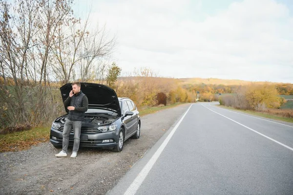 Man repairing a broken car by the road. Man having trouble with his broken car on the highway roadside. Man looking under the car hood. Car breaks down on the autobahn. Roadside assistance concept