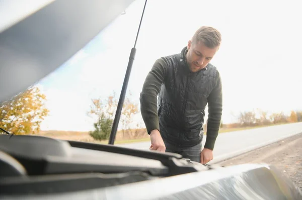 Man repairing a broken car by the road. Man having trouble with his broken car on the highway roadside. Man looking under the car hood. Car breaks down on the autobahn. Roadside assistance concept