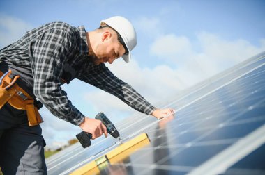 Elinde elektrikli tornavidayla gülümseyen kendinden emin mühendis teknisyeninin portresi bitmemiş yüksek dış güneş paneli voltaik sisteminin önünde duruyor..