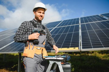 Elinde elektrikli tornavidayla gülümseyen kendinden emin mühendis teknisyeninin portresi bitmemiş yüksek dış güneş paneli voltaik sisteminin önünde duruyor..