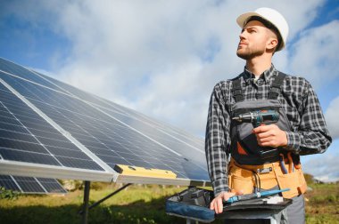 Elinde elektrikli tornavidayla gülümseyen kendinden emin mühendis teknisyeninin portresi bitmemiş yüksek dış güneş paneli voltaik sisteminin önünde duruyor..