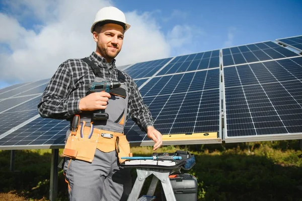 Elinde elektrikli tornavidayla gülümseyen kendinden emin mühendis teknisyeninin portresi bitmemiş yüksek dış güneş paneli voltaik sisteminin önünde duruyor..