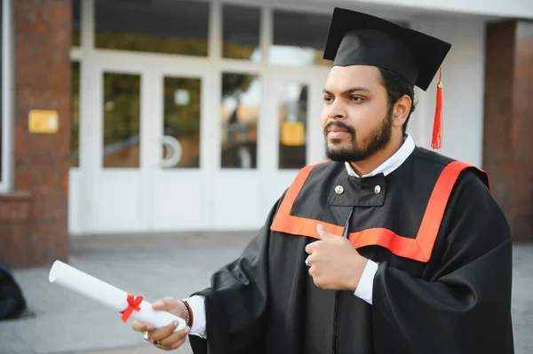 Indian graduate in graduation robe with diploma in university campus copy space
