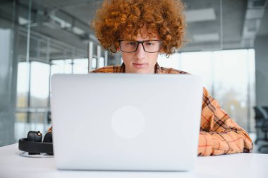 Genç, mutlu, serbest giyinen, dişlek gülümsemeli, kafede dizüstü bilgisayarlı kameraya bakan ve cep telefonu kullanan bir erkek.