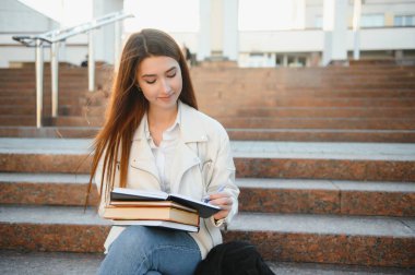 Merdivenlerinde açık havada, kitap okumak oturmak sırt çantası ile neşeli genç kız öğrenci portresi.