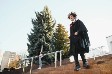 Üniversitenin yanında diplomasını elinde tutan mutlu erkek mezununun düşük açılı portresi. Üniversite mezuniyetini kutlayan akademik başarılardan gurur duyan genç ve yakışıklı bir adamdan..