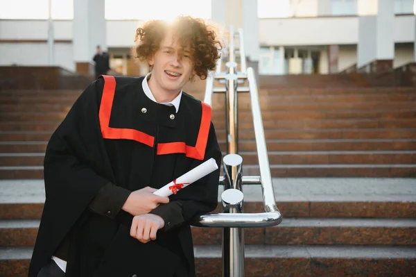 大学卒業 若い笑顔の少年大学卒業生伝統的な骨やマントル立って手に卒業証書を保持大学の建物の背景に — ストック写真