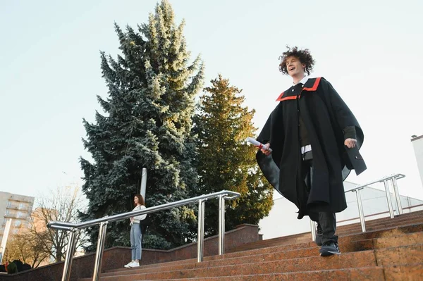 从大学毕业 面带微笑的男孩大学毕业 身披传统的紧身衣 手握大学建筑背景的文凭 — 图库照片