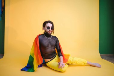 Eşcinsel gurur bayrağı taşıyan eşcinsel adam gülümsüyor ve sarı arka plana bakıyor..