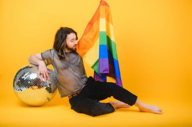 Eşcinsel gurur bayrağı taşıyan eşcinsel adam gülümsüyor ve sarı arka plana bakıyor.