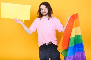 Eşcinsel gurur bayrağı taşıyan eşcinsel adam gülümsüyor ve sarı arka plana bakıyor.