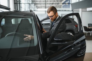 Seçimini yapıyor. Takım elbiseli bir gencin yatay portresi. Arabaya bakıyor ve satın almalı mı diye düşünüyor..