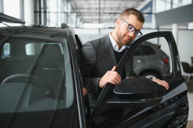 Seçimini yapıyor. Takım elbiseli bir gencin yatay portresi. Arabaya bakıyor ve satın almalı mı diye düşünüyor..