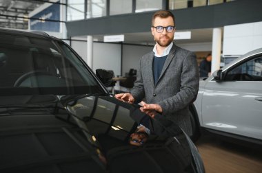 Seçimini yapıyor. Takım elbiseli bir gencin yatay portresi. Arabaya bakıyor ve satın almalı mı diye düşünüyor..