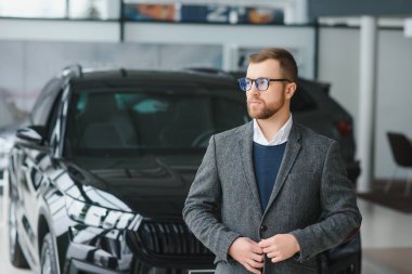 Seçimini yapıyor. Takım elbiseli bir gencin yatay portresi. Arabaya bakıyor ve satın almalı mı diye düşünüyor..