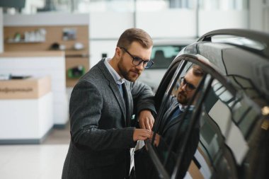 Seçimini yapıyor. Takım elbiseli bir gencin yatay portresi. Arabaya bakıyor ve satın almalı mı diye düşünüyor..