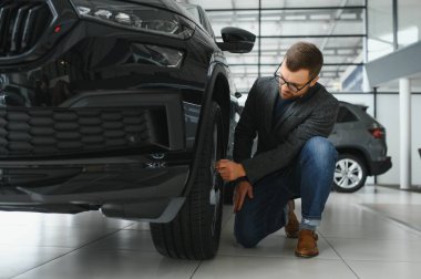 Seçimini yapıyor. Takım elbiseli bir gencin yatay portresi. Arabaya bakıyor ve satın almalı mı diye düşünüyor..