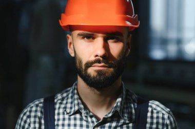 Profesyonel Ağır Sanayi Mühendisi, Güvenlik Üniforması Giyen İşçi, Sert Şapka Gülümseyen. Arka planda Odaklanmamış Büyük Endüstriyel Fabrika