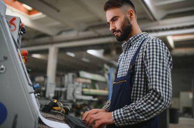 Endüstriyel fabrikada çalışan erkek fabrika işçisi, güvenlik üniforması ve başlık giyerken makineyle birlikte çalışıyor.