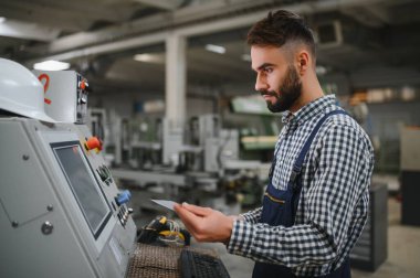 Endüstriyel fabrikada çalışan erkek fabrika işçisi, güvenlik üniforması ve başlık giyerken makineyle birlikte çalışıyor.