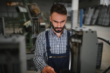 Endüstriyel fabrikada çalışan erkek fabrika işçisi, güvenlik üniforması ve başlık giyerken makineyle birlikte çalışıyor.