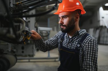 Profesyonel Ağır Sanayi Mühendisi, Güvenlik Üniforması Giyen İşçi, Sert Şapka Gülümseyen. Arka planda Odaklanmamış Büyük Endüstriyel Fabrika