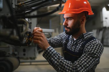 Endüstriyel fabrikada çalışan erkek fabrika işçisi, güvenlik üniforması ve başlık giyerken makineyle birlikte çalışıyor.