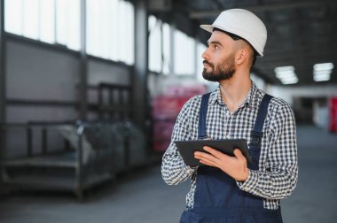Profesyonel Ağır Sanayi Mühendisi, Güvenlik Üniforması Giyen İşçi, Sert Şapka Gülümseyen. Arka planda Odaklanmamış Büyük Endüstriyel Fabrika