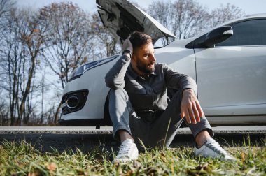 Yolda bozuk bir arabanın yanında duran üzgün şoför kafasını tutuyor. Araba arızası kavramı.