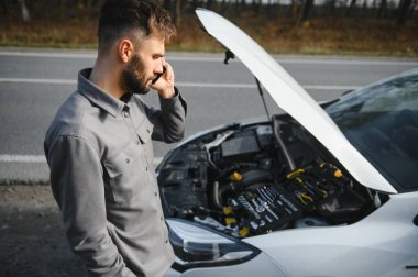 Sad driver calling car service, opening hood, having engine problem standing near broken car on the road. Car breakdown concept.