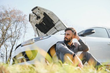 Hayal kırıklığına uğramış adamın yanında kırık araba ile açık örtünün oturan resmi.