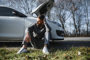 Yolda bozuk bir arabanın yanında duran üzgün şoför kafasını tutuyor. Araba arızası kavramı.